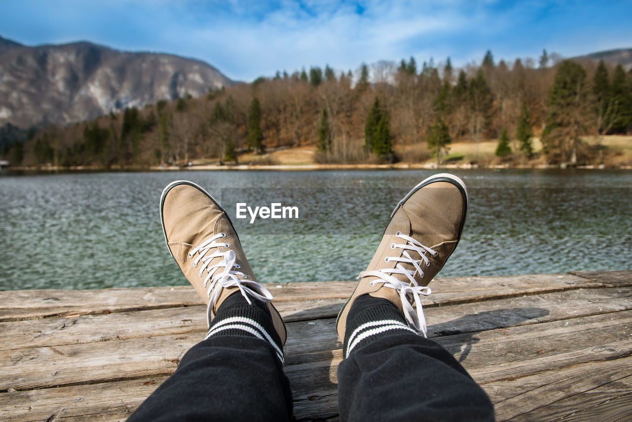 Low section of person in shoes sitting on jetty by lake against sky