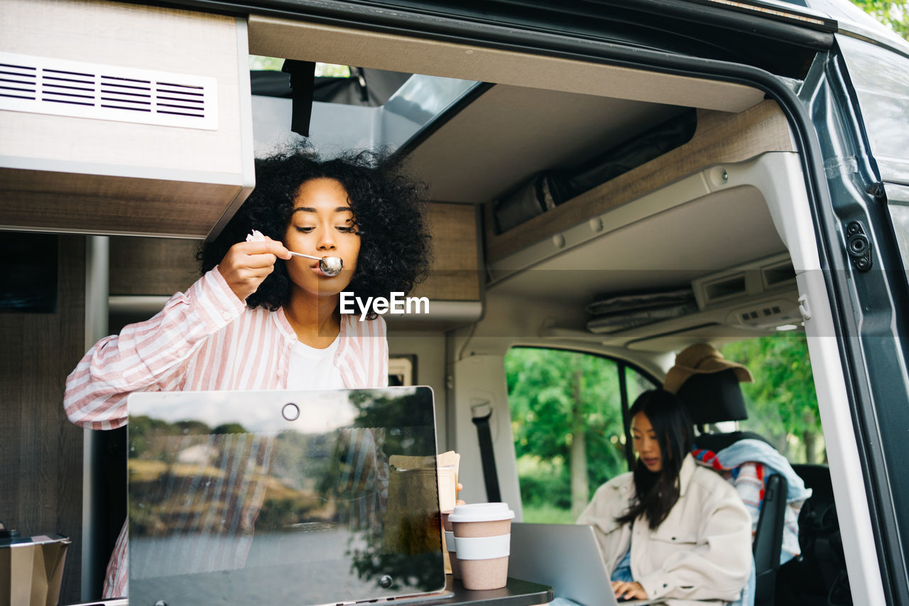 Young black woman preparing coffee while watching video on laptop inside camper vehicle with asian girlfriend during summer journey in nature