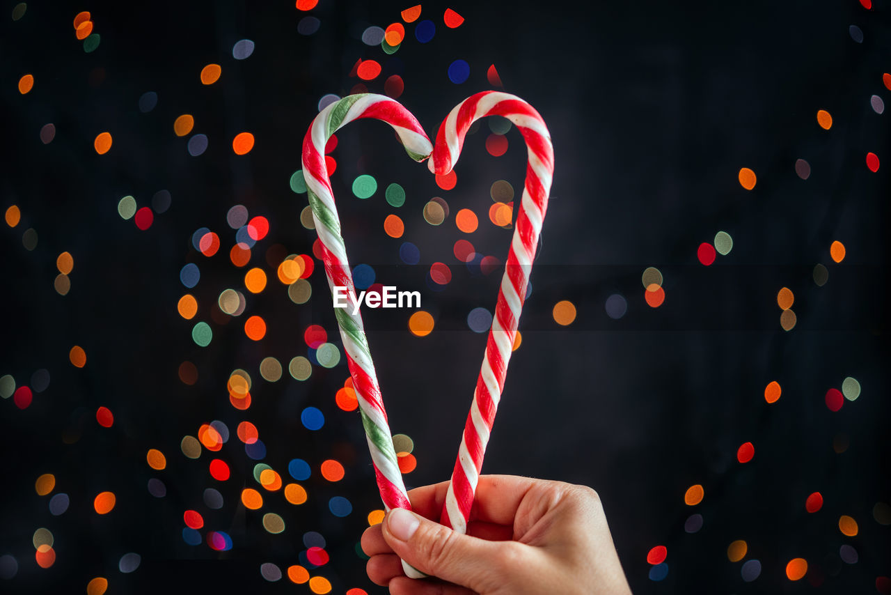 A hand holding candy canes in the shape of heart against christmas tree lights bokeh