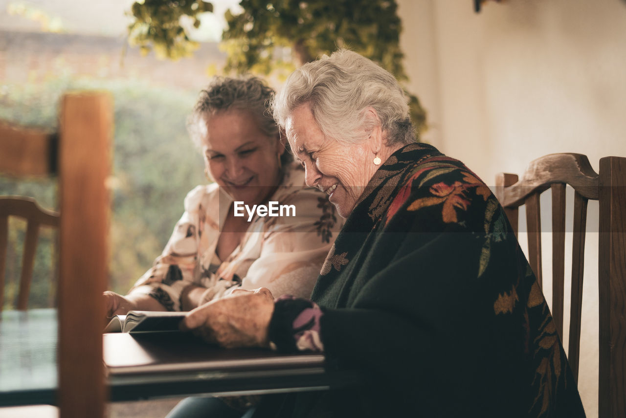 Elderly sisters inspecting pictures in photo album and discussing memories while sitting at table at home together