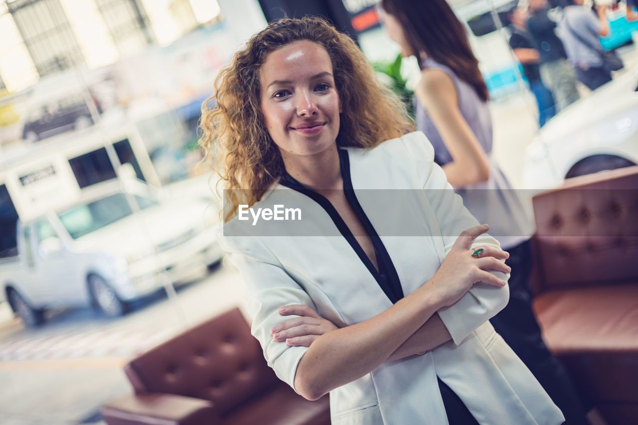 PORTRAIT OF A SMILING YOUNG WOMAN STANDING AGAINST BLURRED BACKGROUND