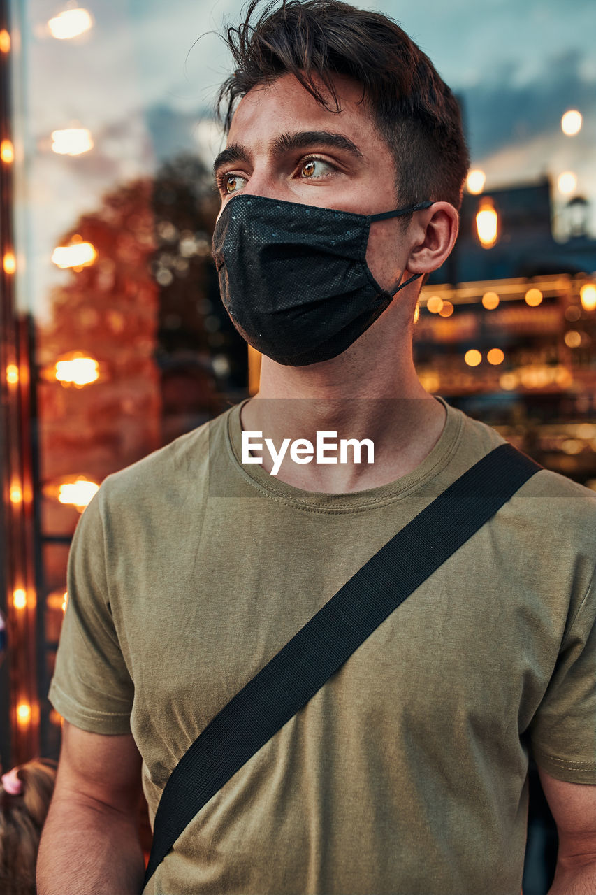 Young man standing at store front in the city center in the evening, looking away