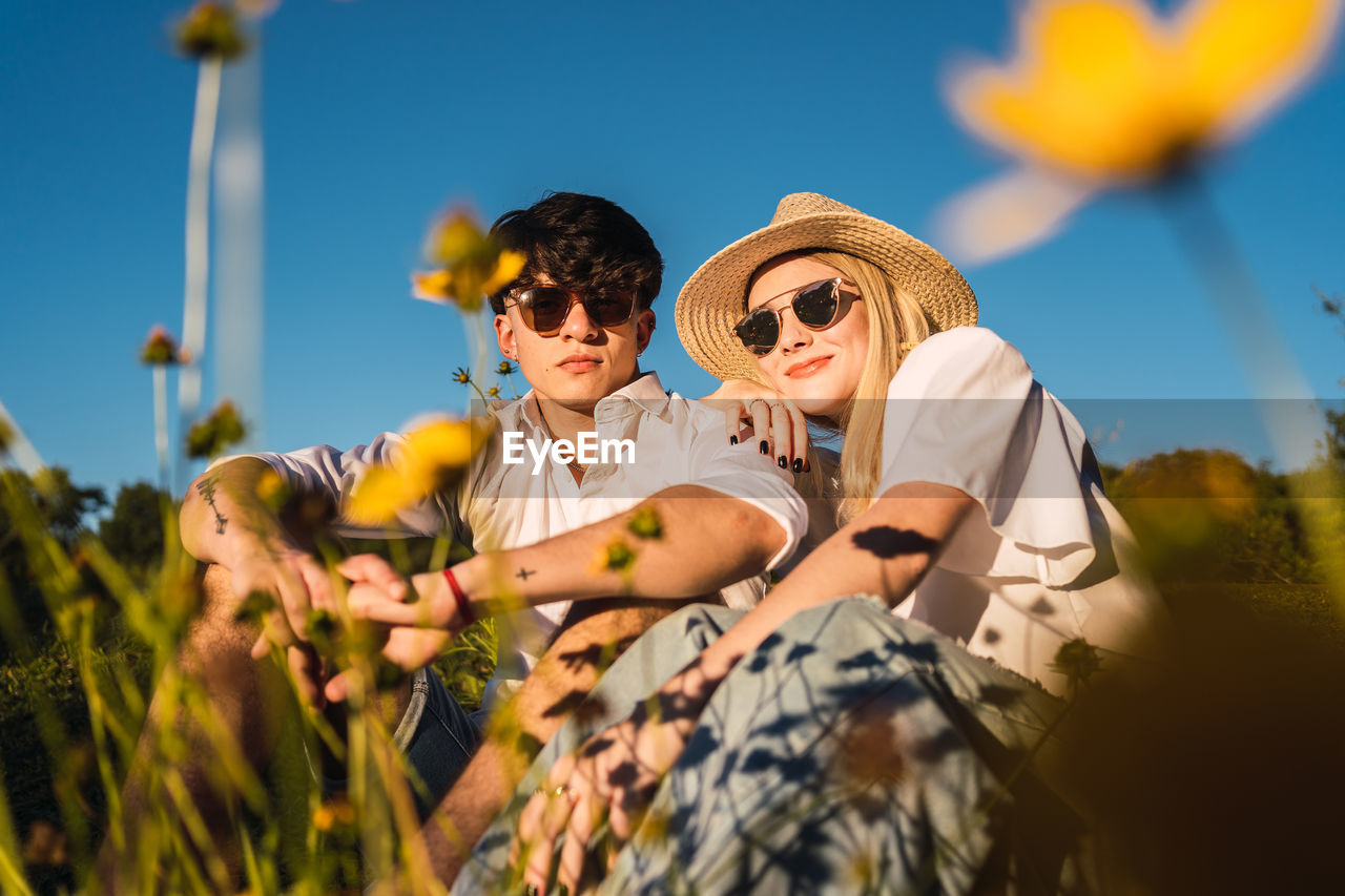 From below cheerful woman and boyfriend in sunglasses smiling and looking at camera while spending time on sunny day in nature