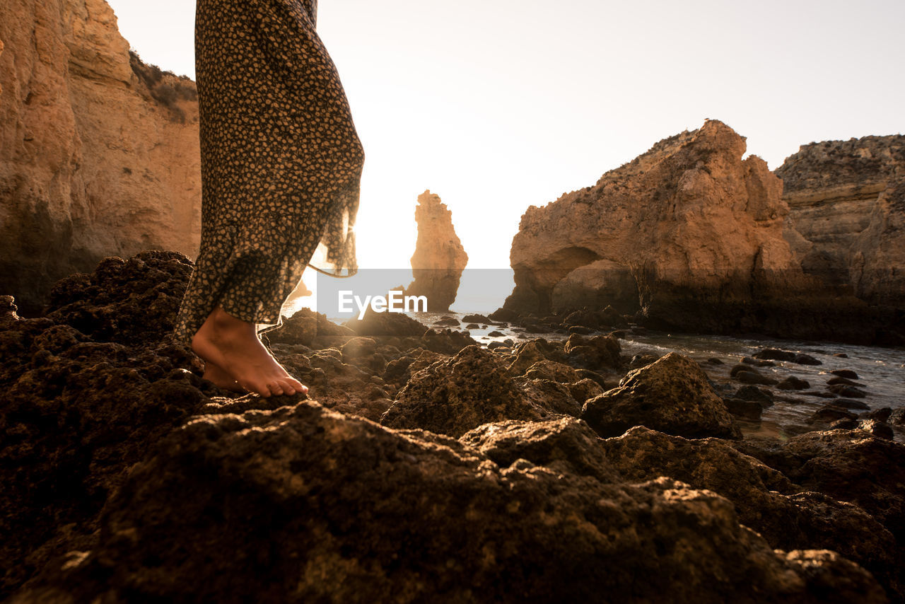 Back view of cropped anonymous female standing barefoot in rocks near sea on sunny day in algar seco caves in algarve, portugal