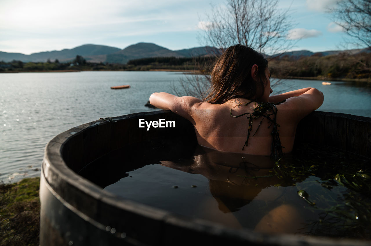 Anonymous woman chilling in barrel with hot water and algae and observing calm lake on weekend day in countryside