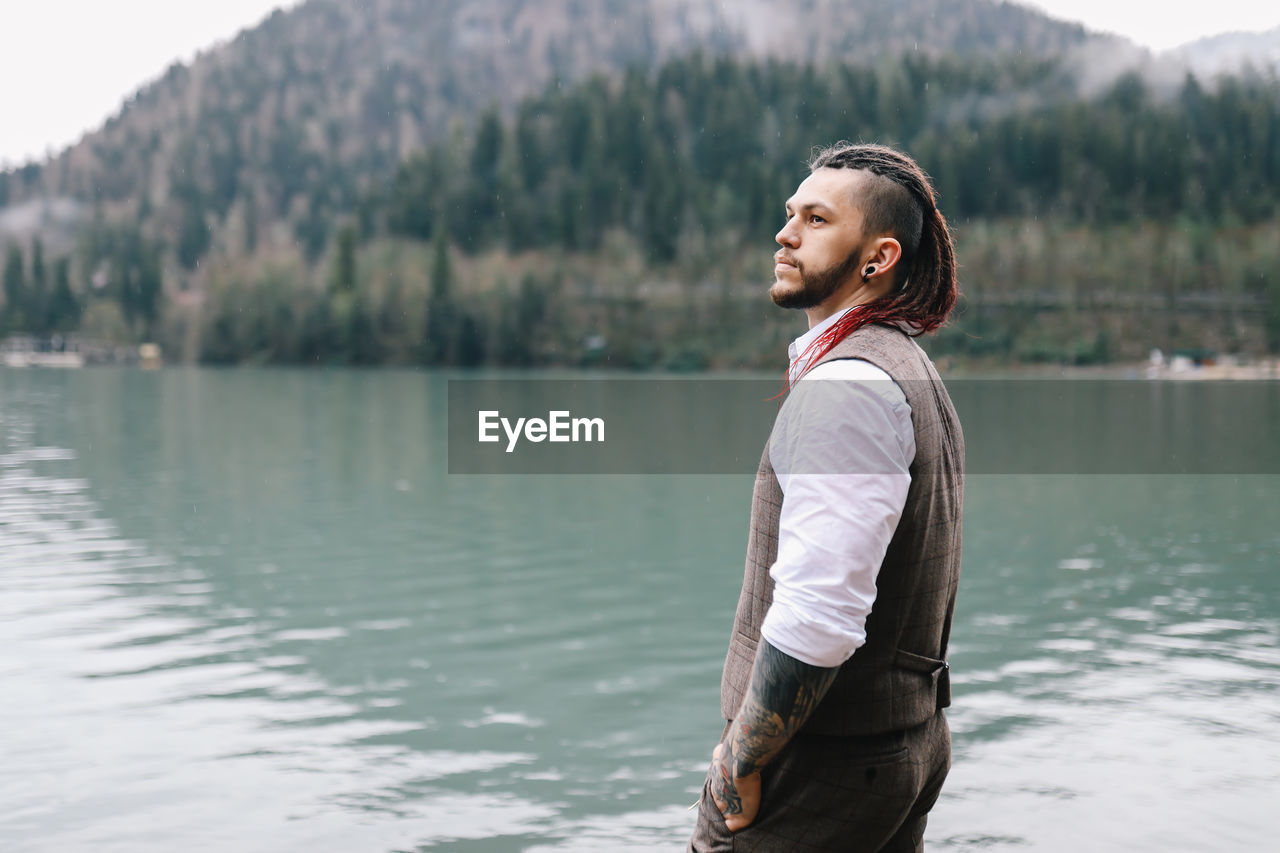 A brutal male hipster groom in a wedding suit in nature by the mountains and lake