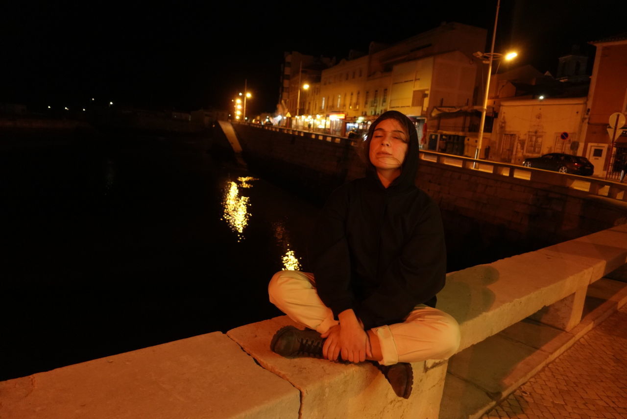 FULL LENGTH OF MAN SITTING AT ILLUMINATED CANAL AT NIGHT