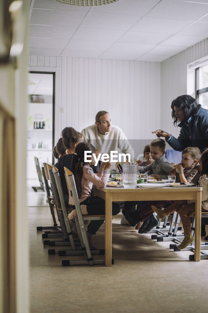 Female and male teachers talking with children having breakfast at dining table