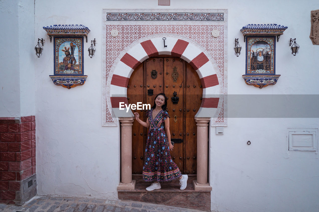 Full body of smiling asian female traveler looking at camera while standing near old ornamental building during trip in spain