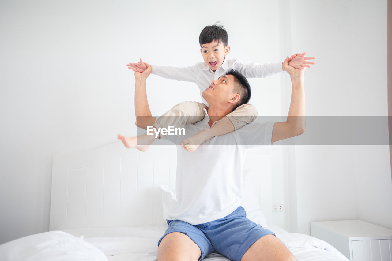 Father carrying son on shoulders while sitting on bed against wall at home