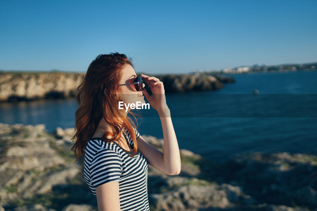 WOMAN WEARING SUNGLASSES STANDING AGAINST SEA AGAINST SKY