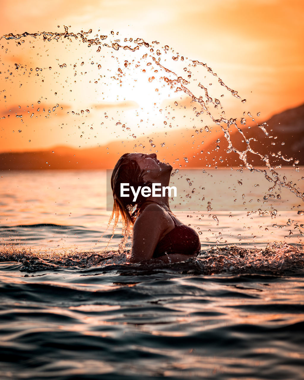 Side view happy fit female in swimwear throwing head back and splashing water while swimming in sea during scenic sunset