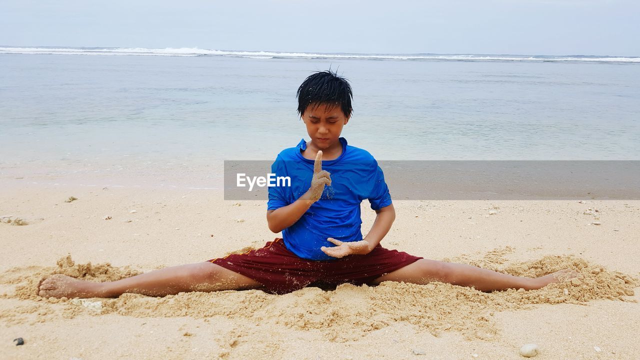 Boy doing the splits on beach against sky