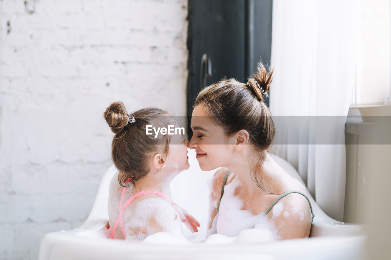 Young mother woman and little tween girl daughter having fun in bath with foam at home