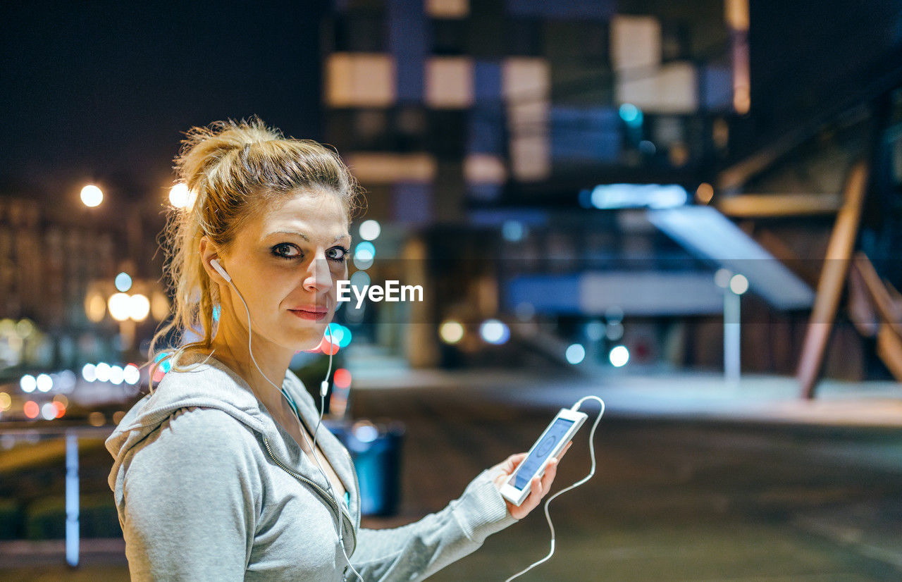 Portrait of young blonde woman looking at camera while listening music on mobile phone application