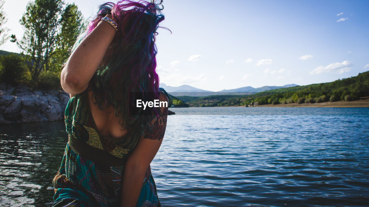 Young woman with hand in hair standing against river against sky