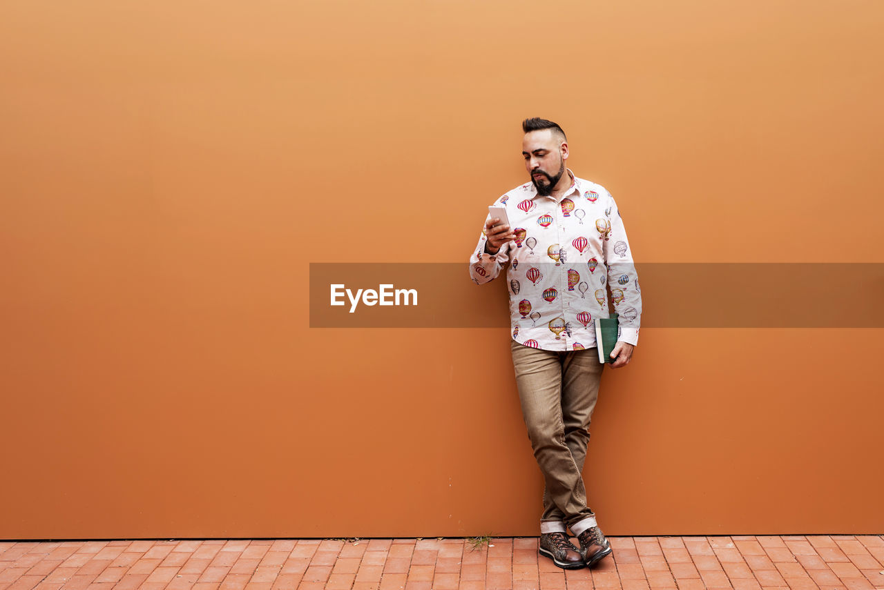 Bearded man leaning on wall while using smartphone by office building