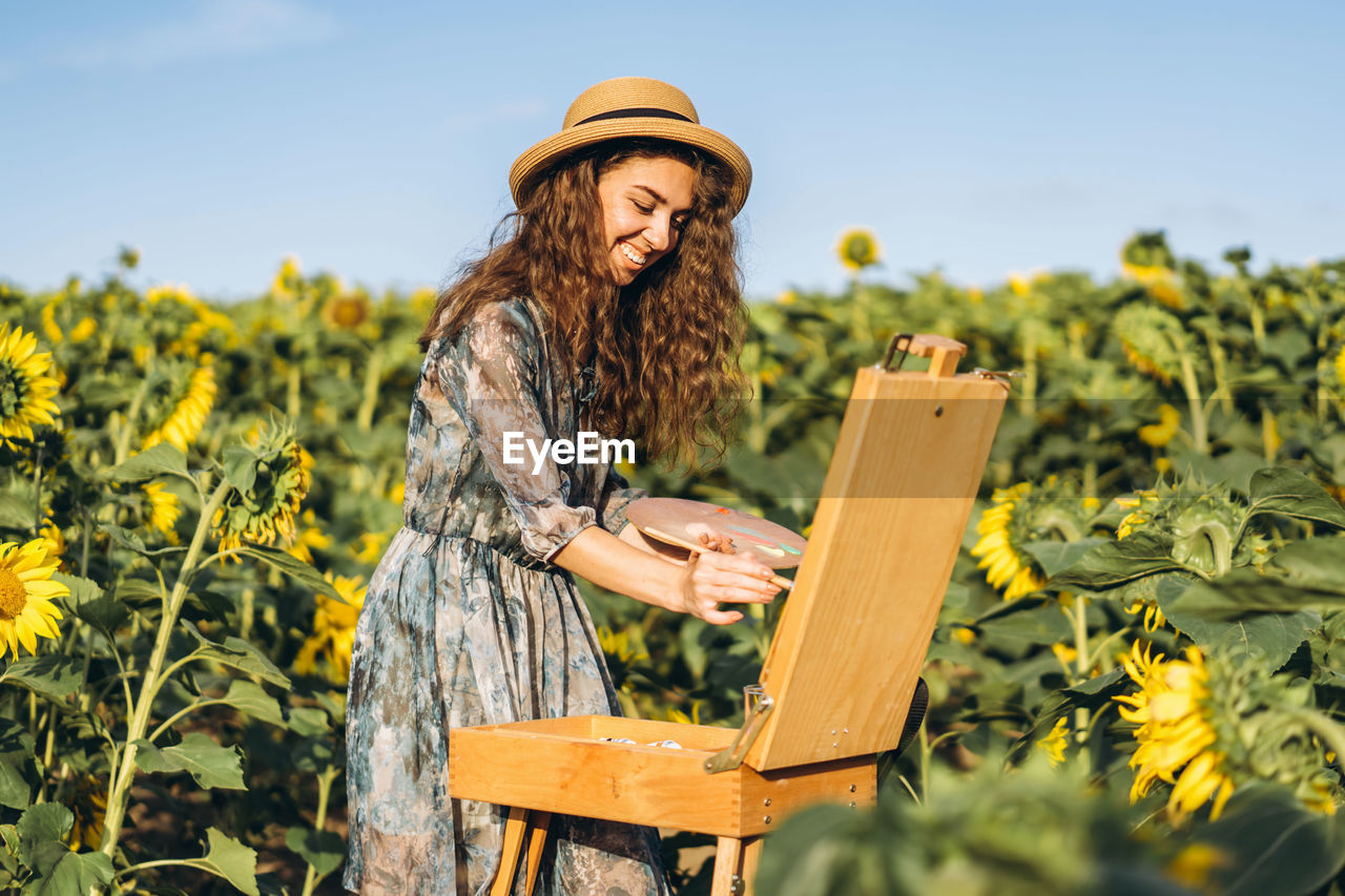 A young woman with curly hair and wearing a hat is painting in nature. 