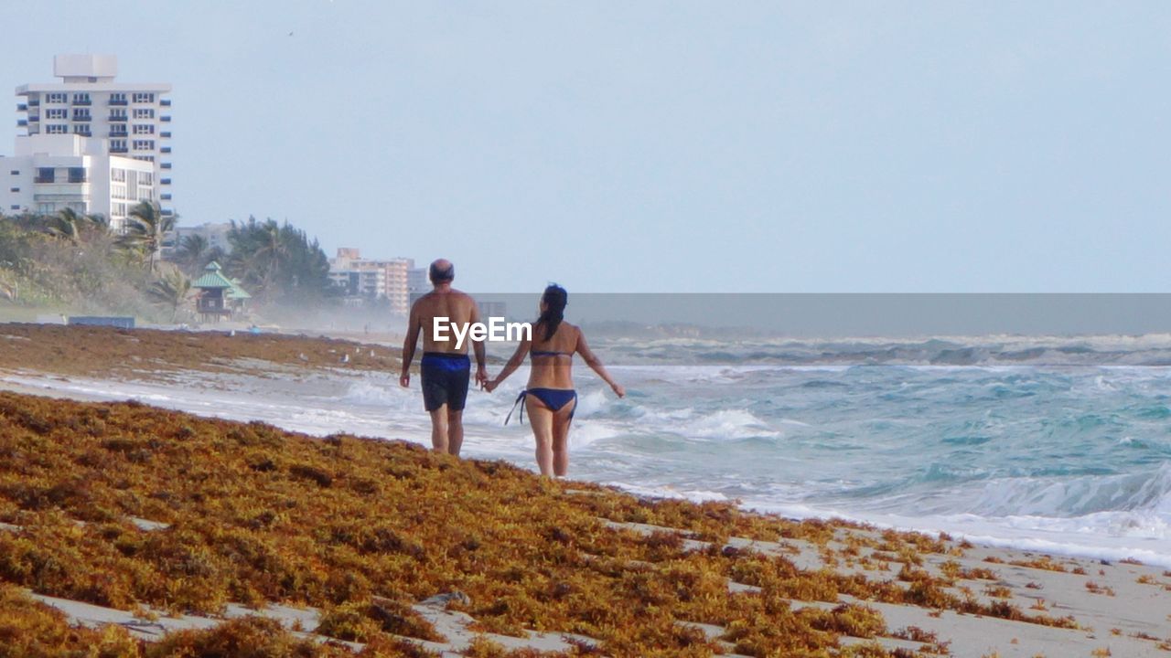 Rear view of couple walking at beach against clear sky