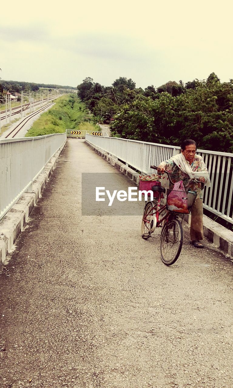 Senior man with bicycle walking on footbridge against sky