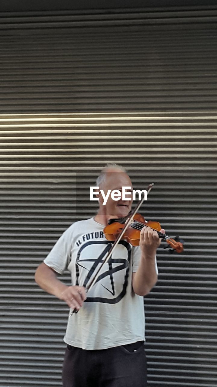 Mature man playing violin while standing against shutter