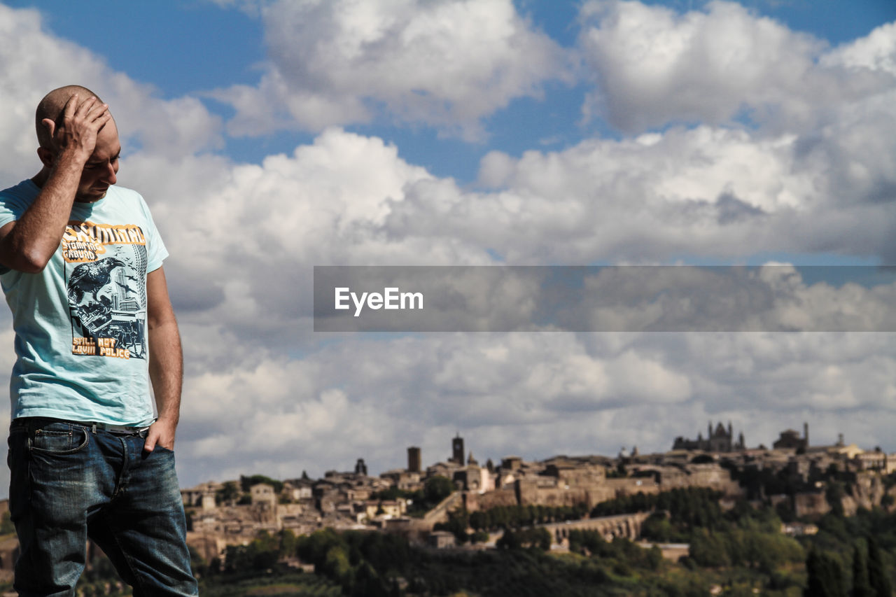 Thoughtful man with hand in head by cityscape against cloudy sky