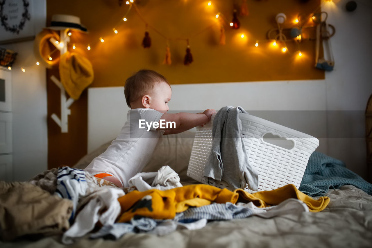 Funny european chubby kid pulling out clothes from laundry basket on bed in cozy bedroom,