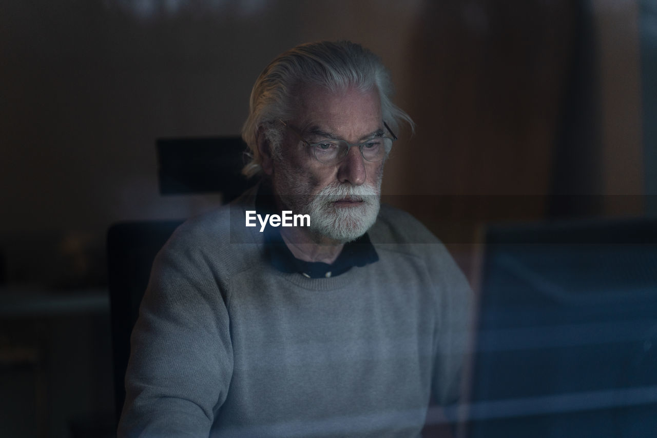 Senior man using computer at home during night seen through glass