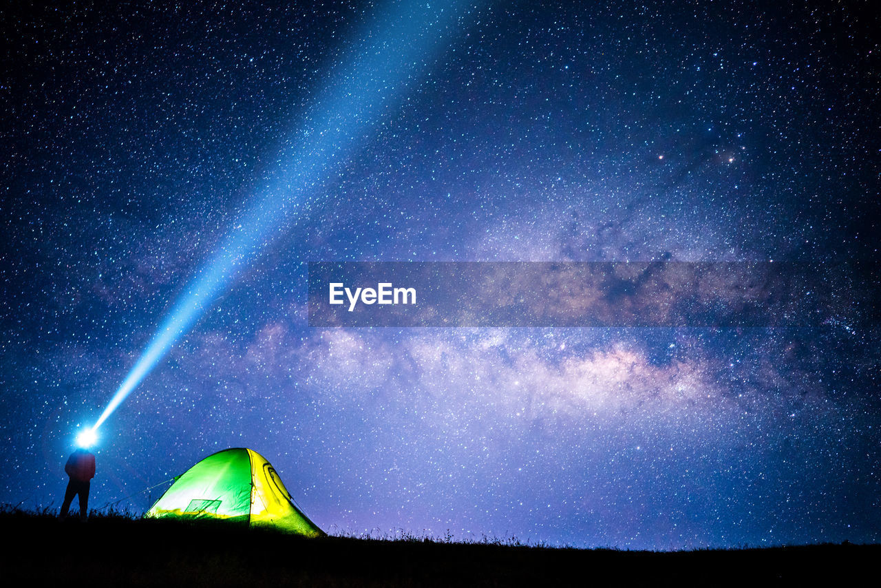 Low angle view of man holding flashlight while standing by tent on field against sky