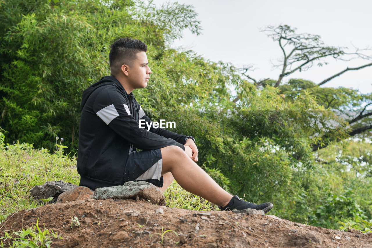 one person, plant, sitting, tree, nature, leisure activity, lifestyles, full length, young adult, casual clothing, adult, looking, side view, men, relaxation, contemplation, looking away, day, outdoors, land, person, clothing, sky, green, forest, rock