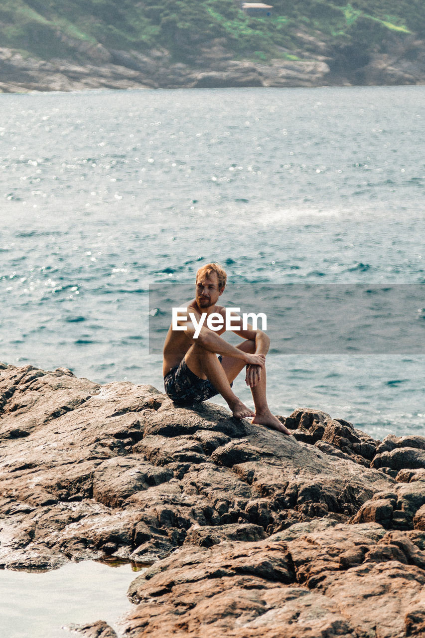 Full length of shirtless man sitting on rock at beach