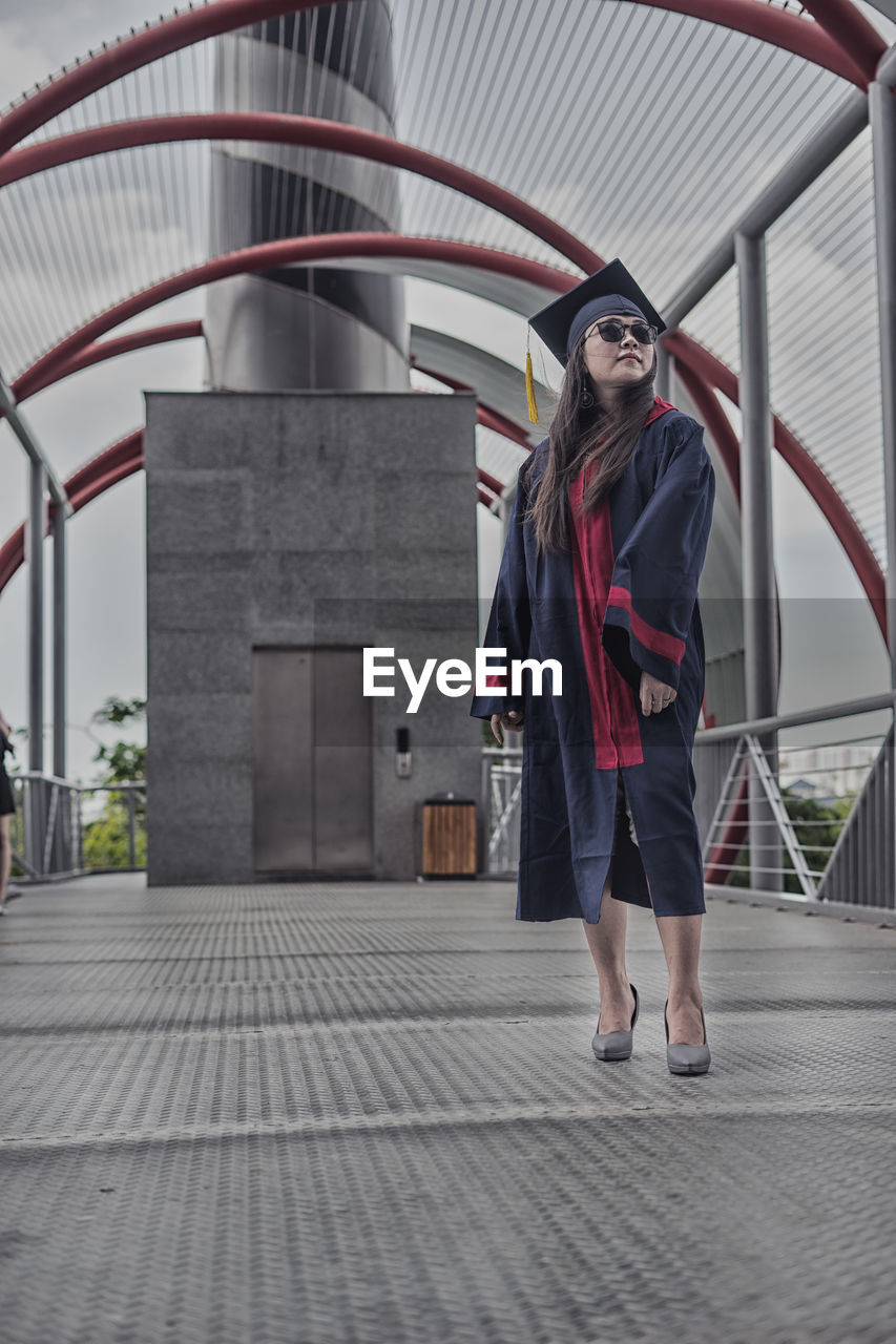 Low angle view of young woman wearing graduation gown while standing on footbridge