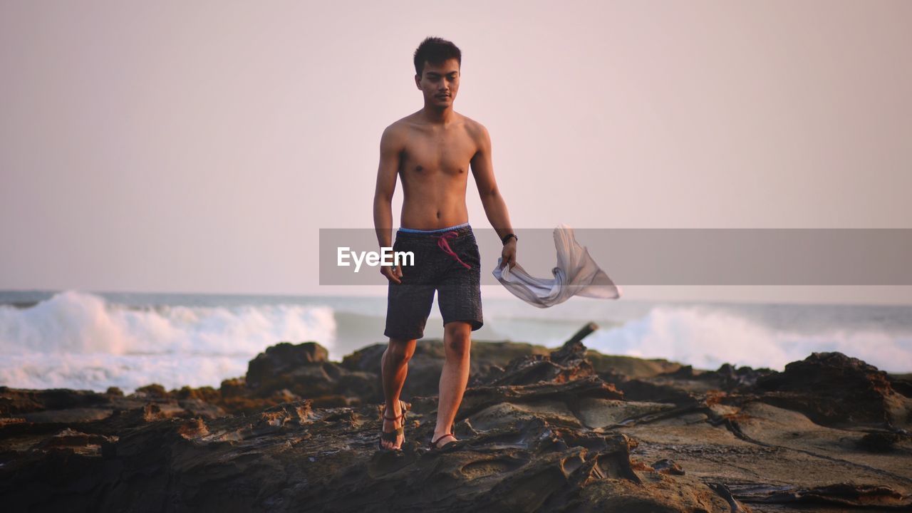 Shirtless mid adult man with tank top walking at beach against clear sky