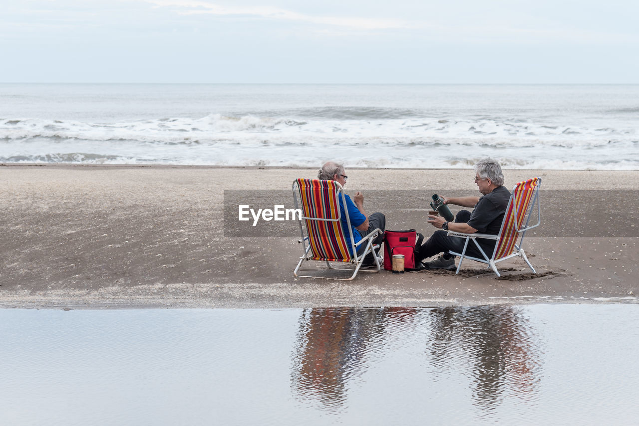 Two mature gray haired men sitting on beach chairs pouring water into a mate. 