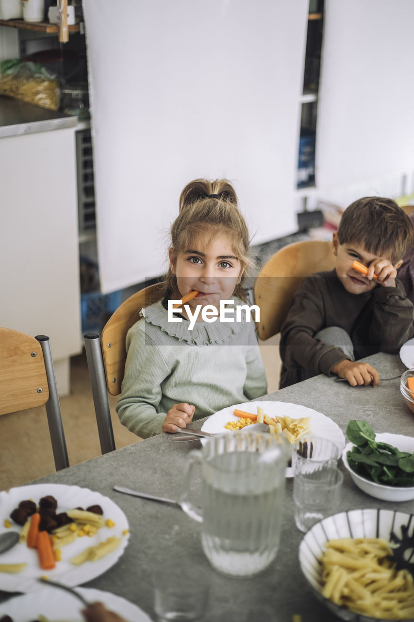 Portrait of playful girl holding carrot slice in mouth while sitting at dining table during lunch time