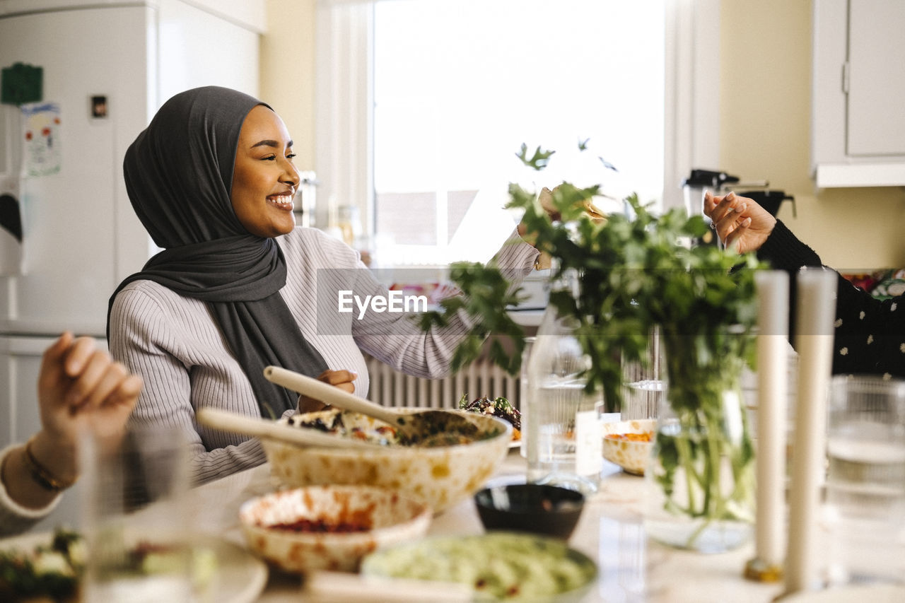 Happy young woman wearing hijab passing food to friend at dining table