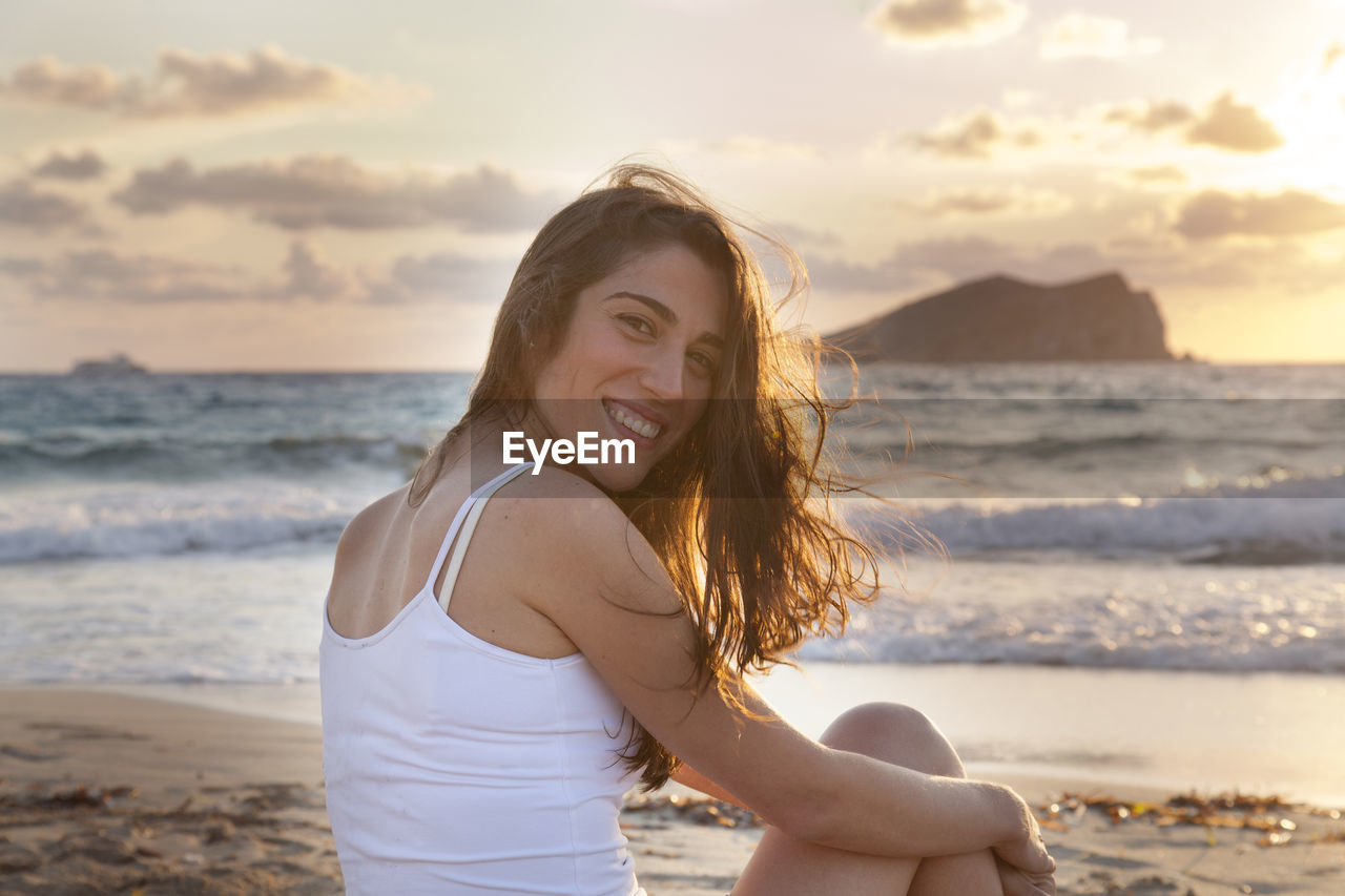 PORTRAIT OF SMILING YOUNG WOMAN STANDING AT BEACH AGAINST SKY