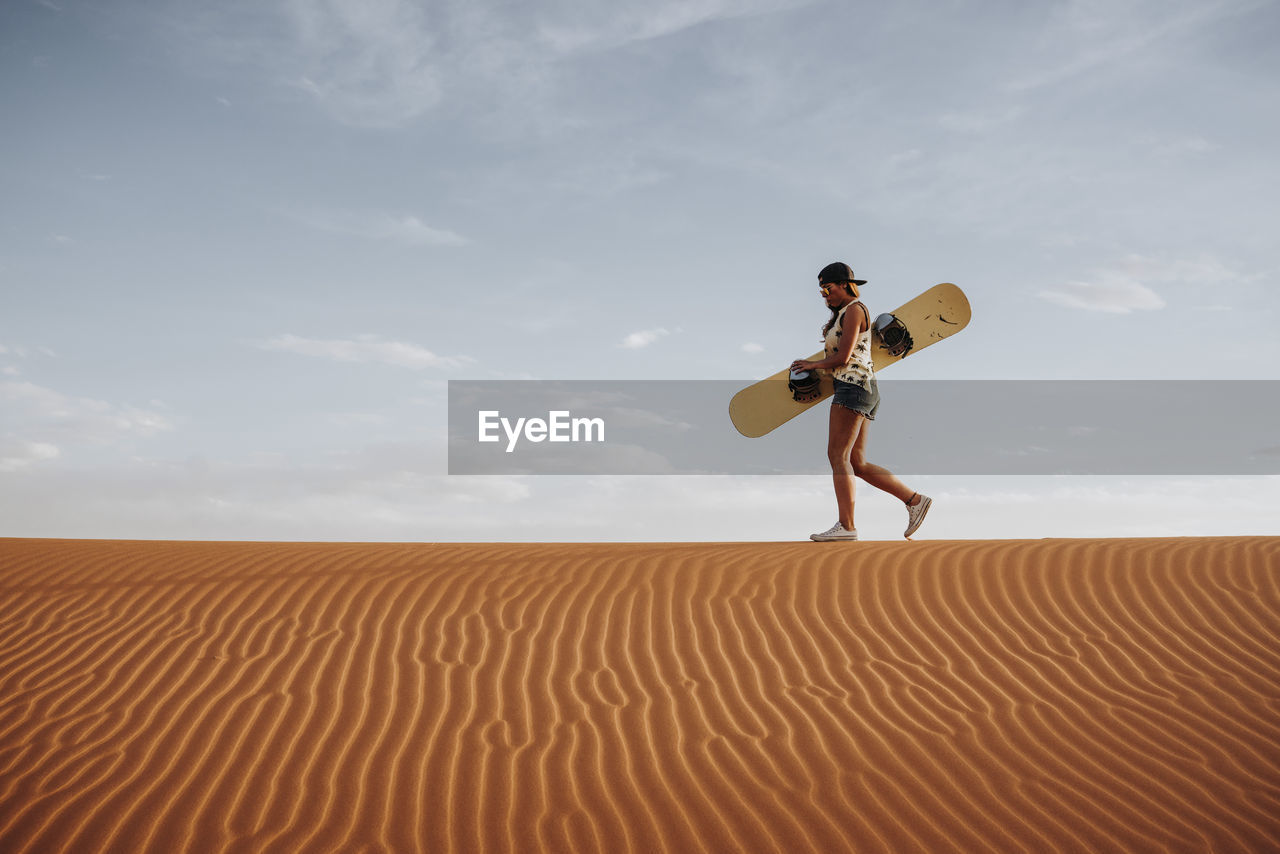 Side view of young female walking on sand and preparing for sandboarding looking away