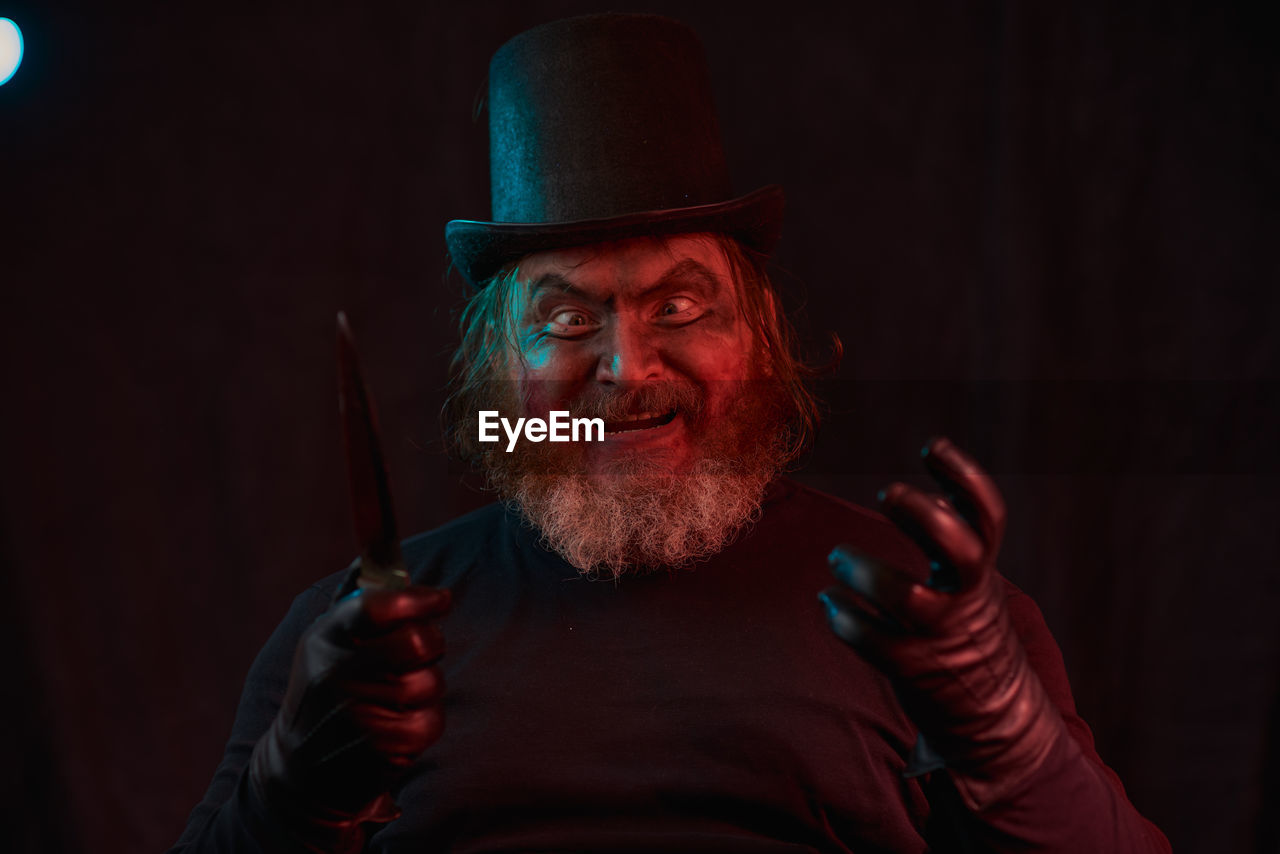 facial hair, adult, beard, one person, portrait, hat, men, performing arts, clothing, red, fashion accessory, darkness, human face, emotion, person, dark, fedora, studio shot, night, indoors, looking at camera, front view, headshot, entertainment, performance, black background, evil, celebration, senior adult