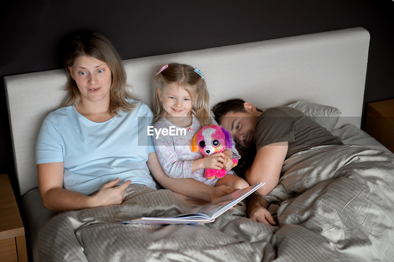 High angle view of mother reading book for daughter while father sleeping on bed at home