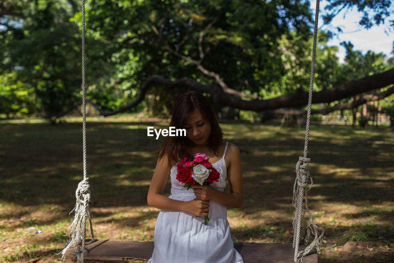 Young woman holding roses sitting on swing at land