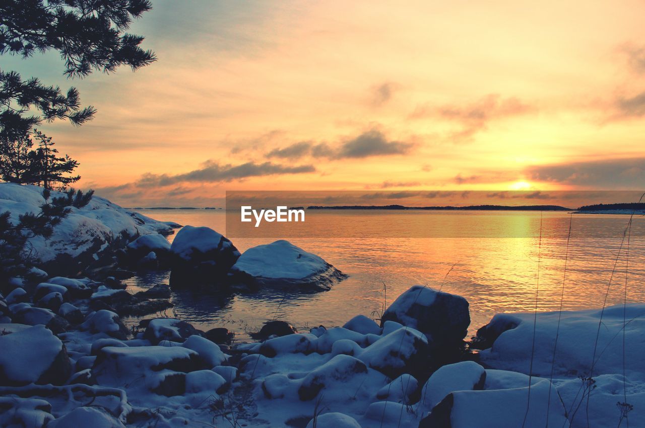 Snow covered riverbank against sky during sunset