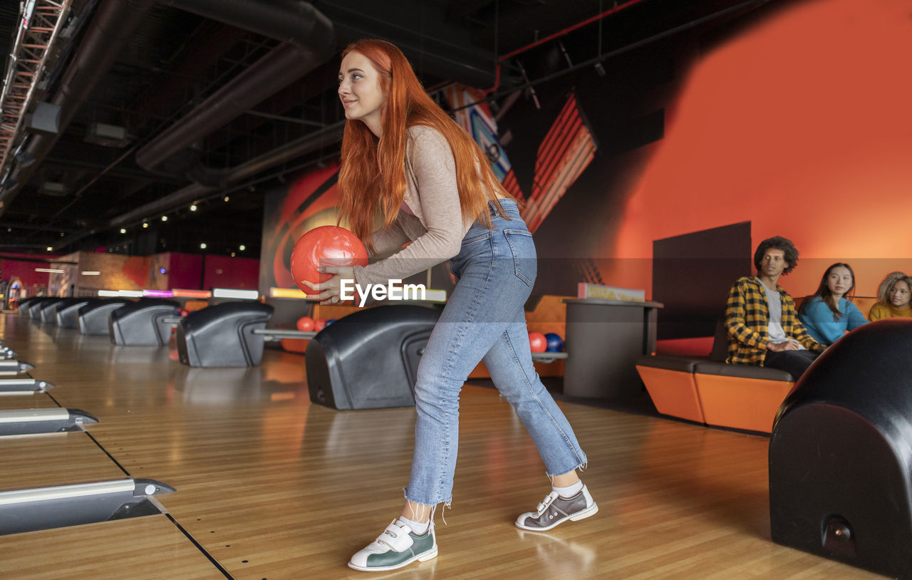 Young woman throwing ball with friends sitting in background at bowling alley