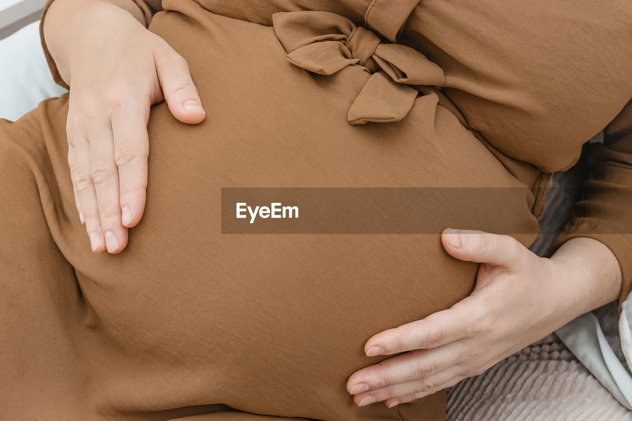 A pregnant woman is holding her belly with her hands, lying on the bed, top view, close-up.