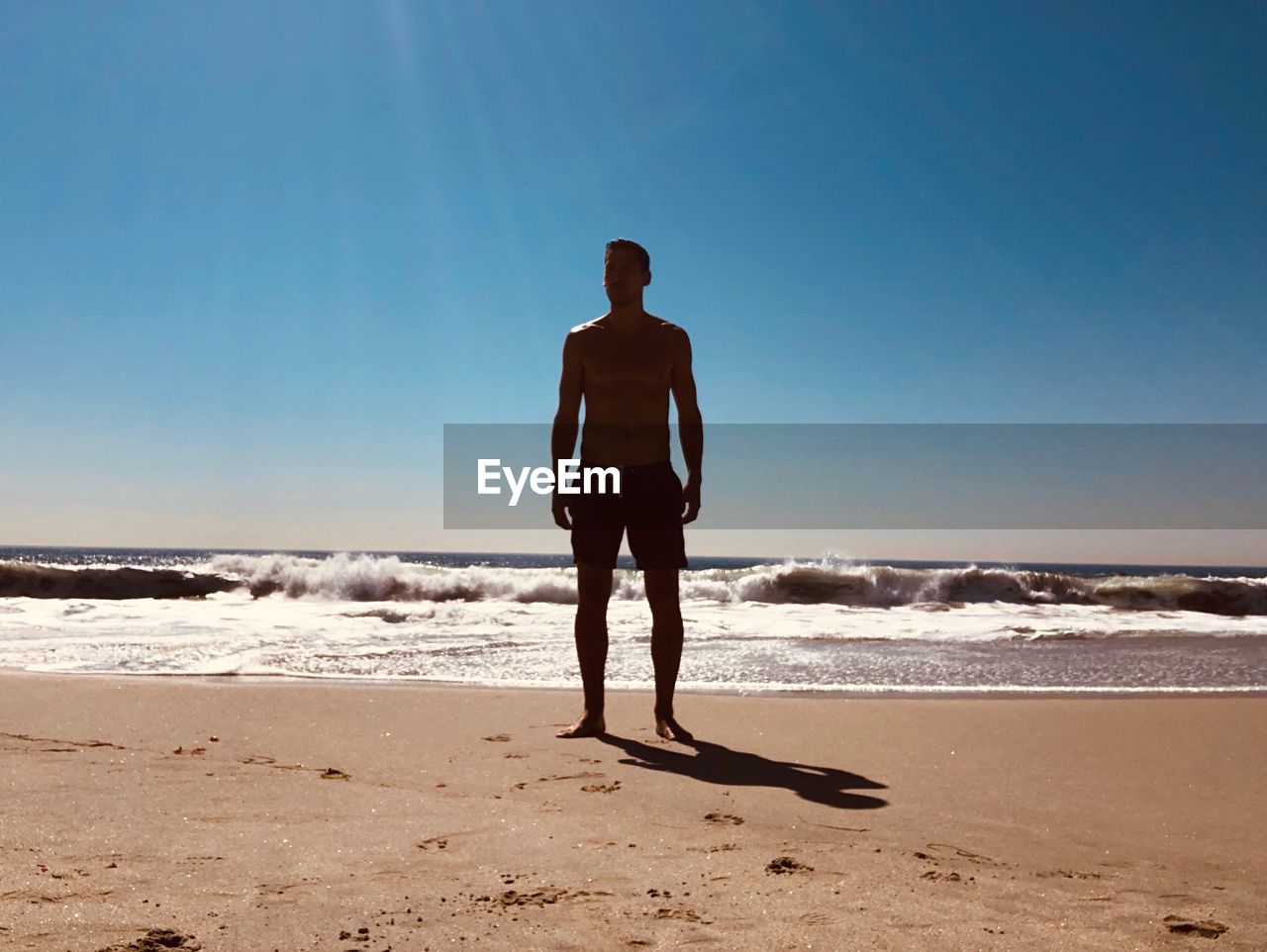 REAR VIEW OF MAN STANDING ON BEACH AGAINST SKY