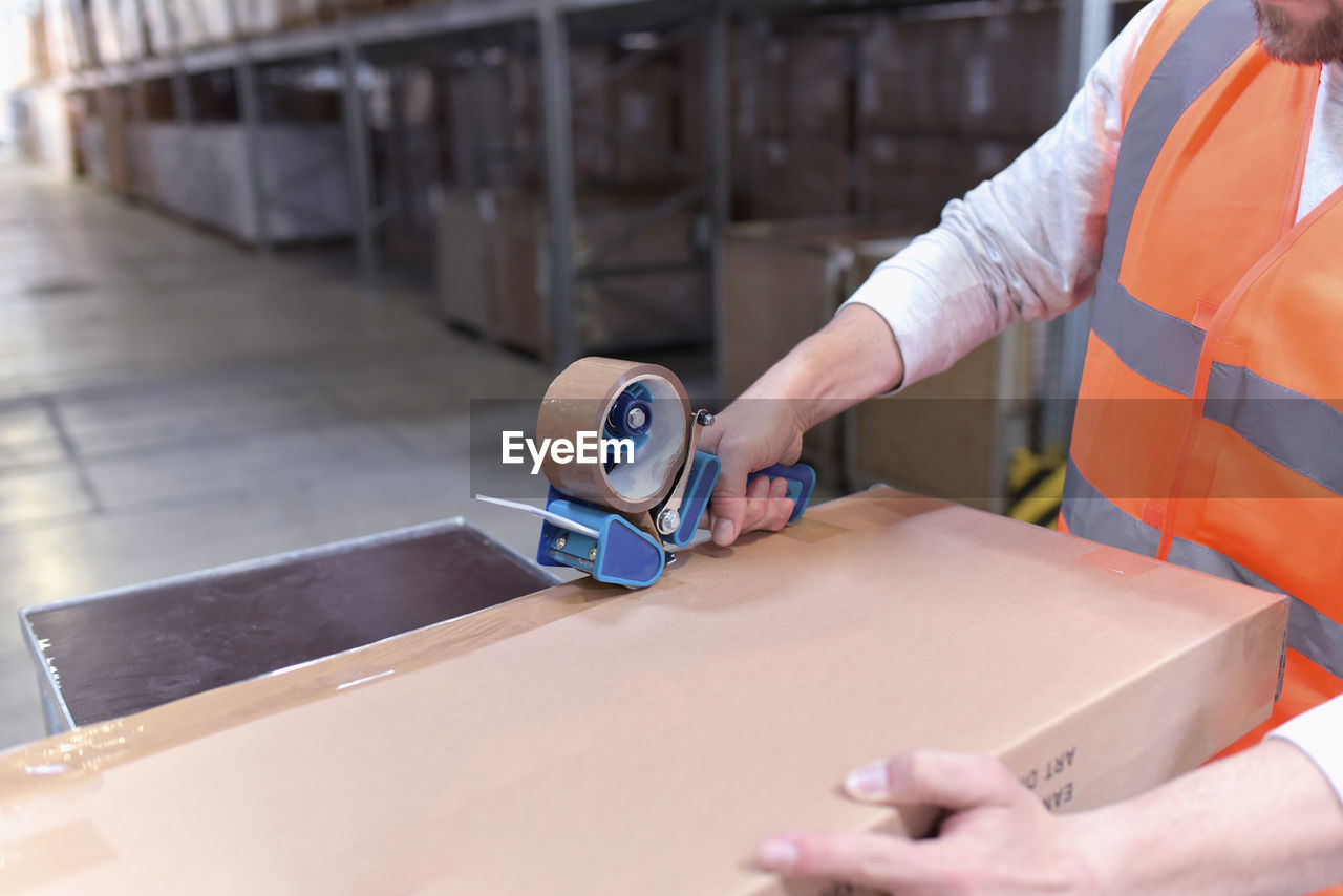 Man in factory hall wearing safety vest closing cardboard boax