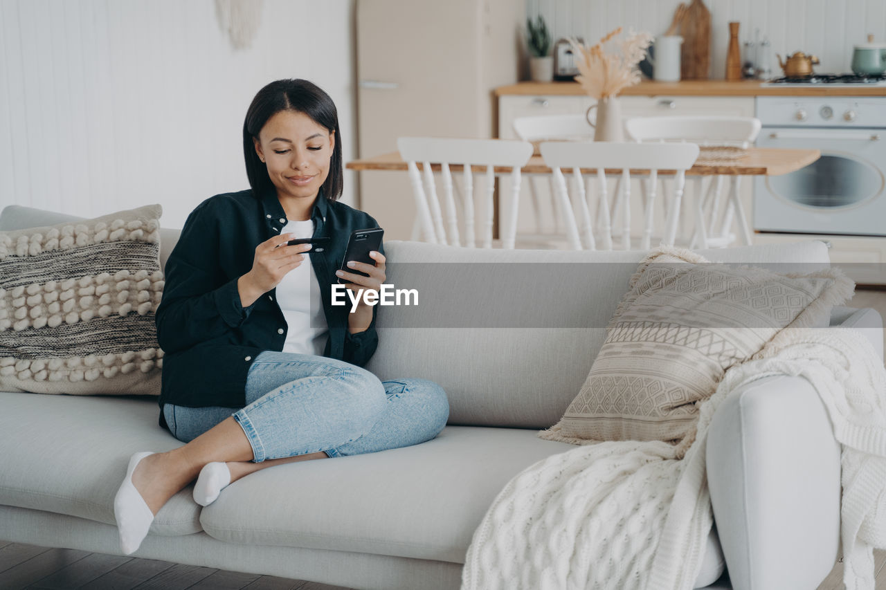 portrait of young woman using phone while sitting on sofa at home