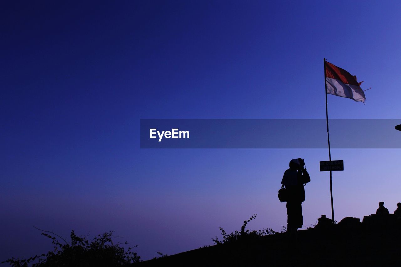 Silhouette man photographing while standing by flag against clear sky