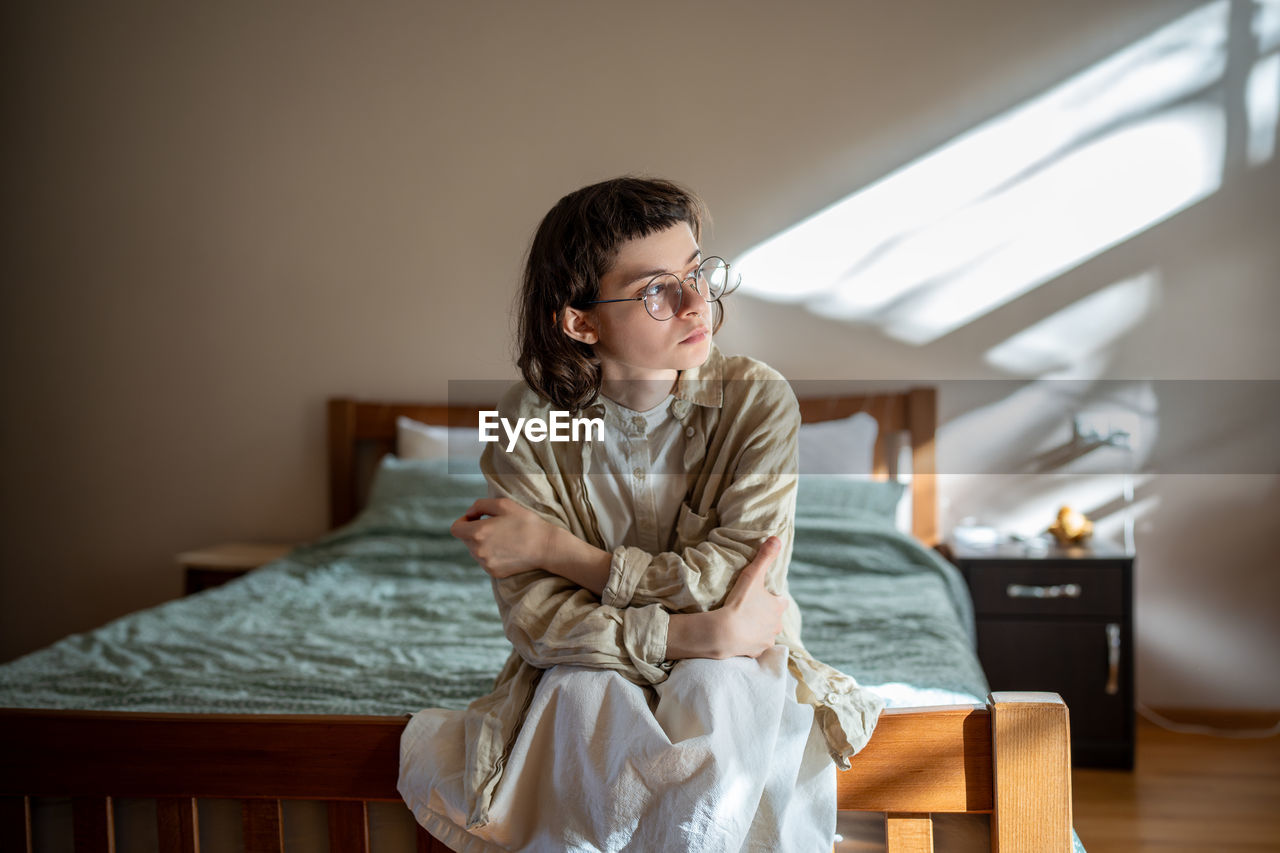 Pensive teen sitting on bed, looking at window, thinking of misunderstanding with friends, parents