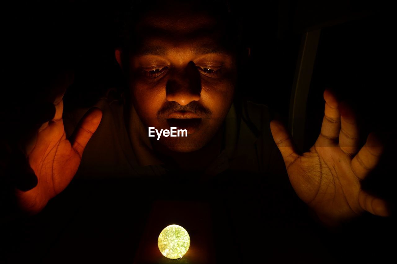 Close-up of man looking at illuminated ball in darkroom
